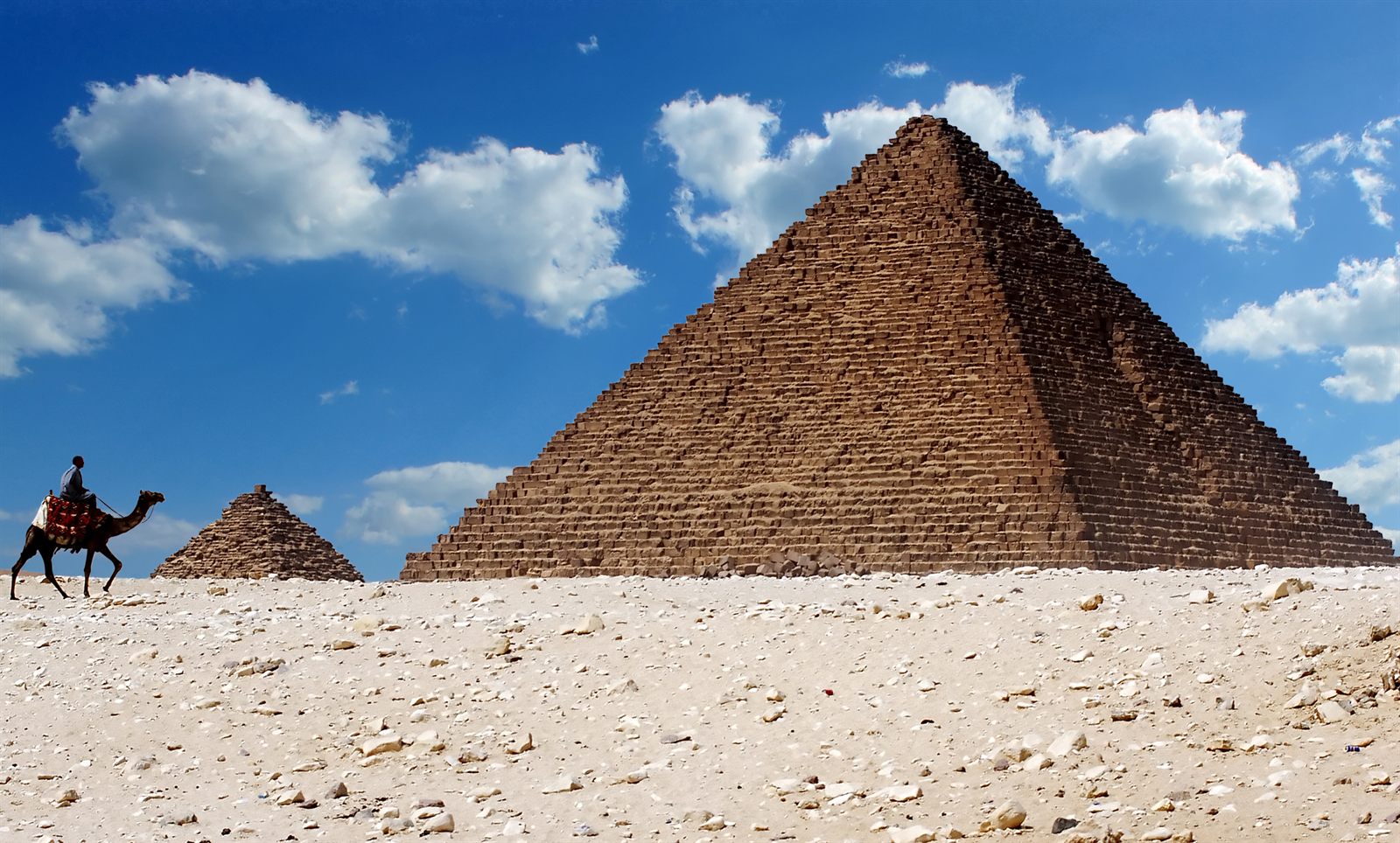 A Giza pyramid under a blue sky with a camel rider crossing the sand